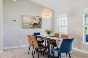 Un coin repas avec une table dressée pour quatre personnes, entourée de chaises aux couleurs variées, un luminaire suspendu et un tableau accroché au mur.