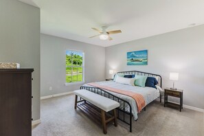 Een slaapkamer met een bed, een bankje, nachtkastjes, een plafondventilator, een raam met uitzicht en een schilderij aan de muur.
