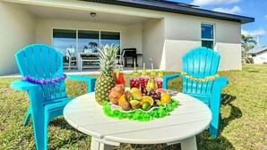 Une terrasse avec deux chaises bleues, une table ronde sur laquelle se trouve un panier de fruits, et une maison en arrière-plan.