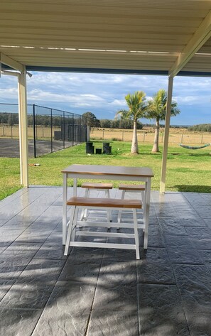 A covered outdoor seating area with a bench, a table, and a view of a grassy field with trees and a fence in the background. - Beautiful Large Country Home in the Perfect Location with tennis court & theatre (Worrigee)