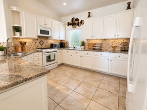 A kitchen with white cabinets, a granite countertop, a microwave, stove, and a refrigerator. - Escape to your Desert Hideaway!  (Thousand Palms)