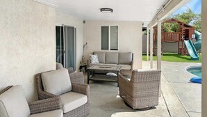 En overdækket terrasse med fletningsmøbler, en polstret sofa, et sofabord og en trælegeplads med rutchebane.