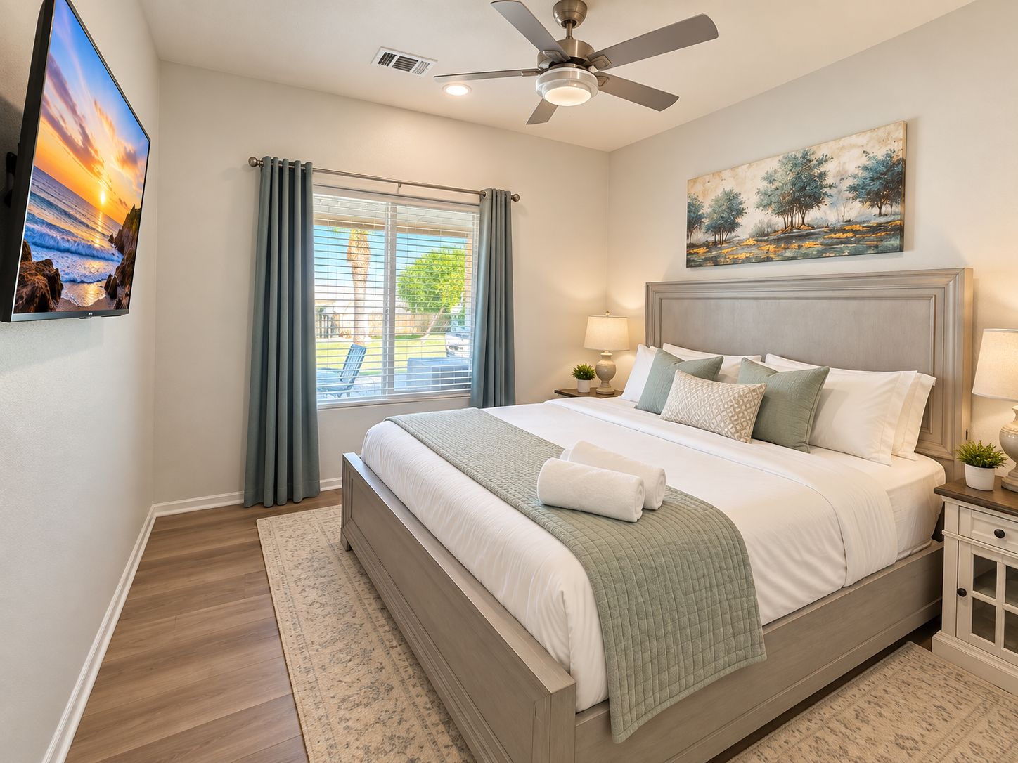 A bedroom with a bed, a ceiling fan, a window with curtains, a painting, and a nightstand with a lamp.