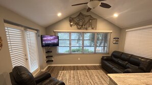 A living room with a black leather sofa, a black leather chair, a flat-screen TV mounted on the wall, a ceiling fan, and a window with blinds. - Lakehouse Living w/ Firepit and Boat Dock to relax and Enjoy the Sunset (palm harbor)