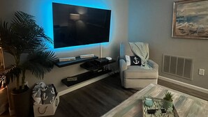 A living room with a flat-screen TV mounted on the wall, a comfortable armchair, a coffee table, and a potted plant. - Lakehouse Living w/ Firepit and Boat Dock to relax and Enjoy the Sunset (palm harbor)