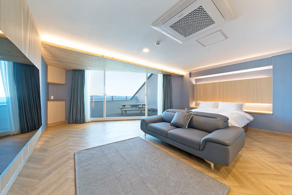 A modern hotel room with a sofa, a bed, a large window, and a view of the outdoors.