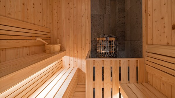 A wooden sauna with benches, a stone wall, and a metal container with rocks. - Oceanterra (Geoje)