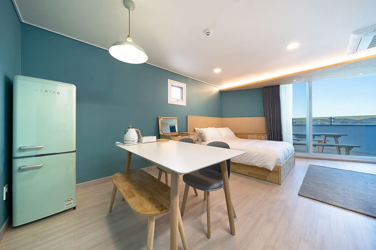A modern hotel room with a bed, a dining table, a small kitchenette, and a large window offering a view of the sea.