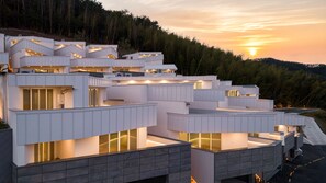 A modern multi-story building with a series of balconies and large windows, set against a backdrop of a forested hillside during sunset.