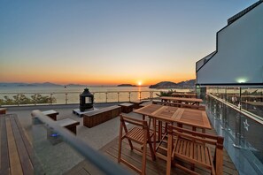 A terrace with wooden furniture and a view of the sunset over the water.