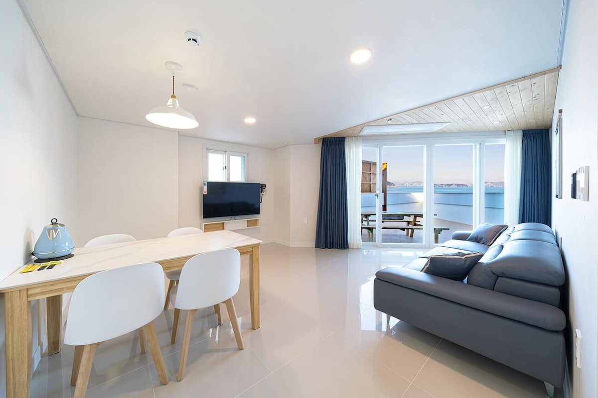 A modern living room with a sofa, a dining table with chairs, a flat-screen TV, and a large window with a view of the sea.