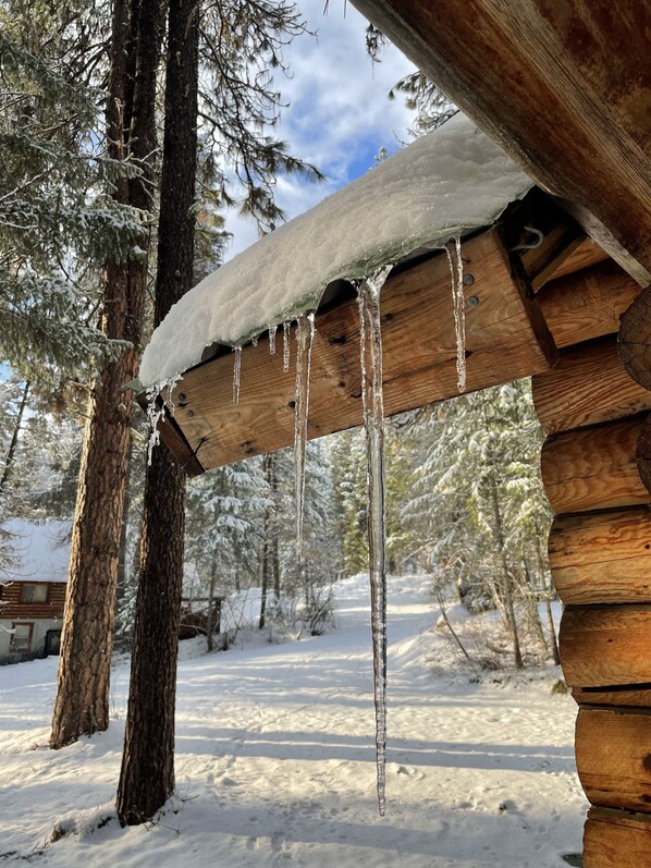 Une cabane en bois, avec des glaçons pendants du toit, au cœur d’une forêt enneigée.