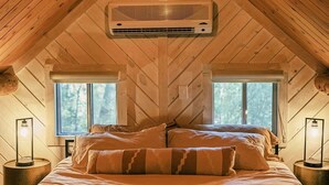 Ein gemütliches Schlafzimmer im Dachgeschoss mit schrägen Wänden, einem großen Bett mit gemusterten Kissen, zwei Nachttischen mit Lampen und einer zentralen Klimaanlage.