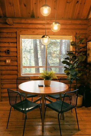 L’intérieur d’un chalet en bois comprend une table à manger ronde, six chaises, deux luminaires sphériques suspendus, une grande fenêtre et une plante en pot.