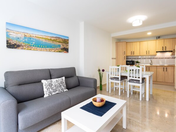 Una sala moderna con un sofá gris, una mesa de centro blanca con un cuenco, y un cuadro de un pueblo costero.