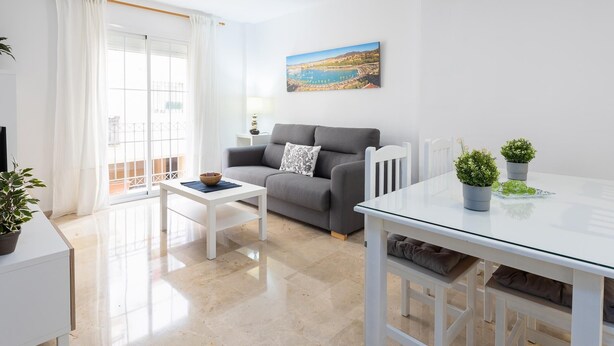 Un salon moderne avec un canapé gris, une table basse en verre et une table à manger avec des chaises. Un tableau orne le mur et une plante en pot est posée sur la table.
