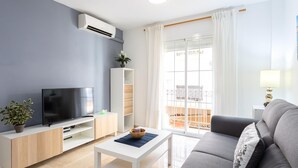 Un salon moderne doté d’un téléviseur à écran plat, d’une table basse blanche, d’un canapé gris et d’un balcon agrémenté de rideaux blancs.
