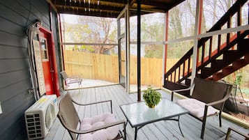 Eine überdachte Terrasse mit Holzdeck, einem Glastisch, zwei Stühlen, einer kleinen Pflanze und einer roten Tür mit der Aufschrift 'GUEST'.