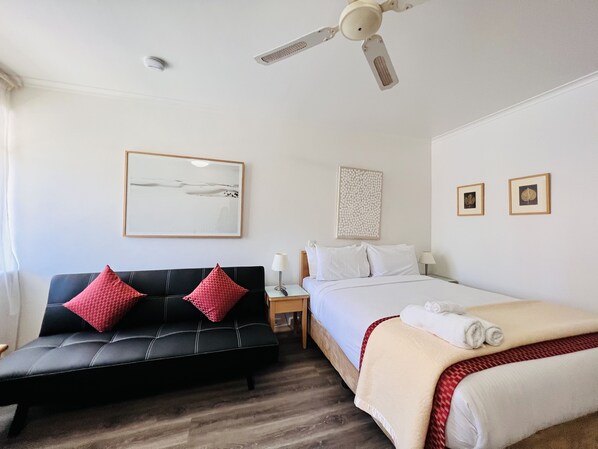 Habitación de hotel con un sofá negro, una cama, un ventilador de techo, una mesita de noche y dos cuadros en la pared.