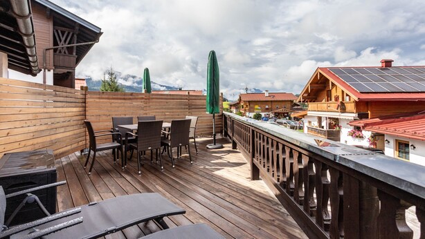 Eine Holzterrasse mit Sitzgelegenheiten im Freien, einem Geländer und Blick auf Gebäude und Berge.