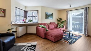 Ein Wohnzimmer mit einem roten Sofa, einem schwarzen Sessel, einem kleinen Tisch, einer Heizung, einem Fenster mit Vorhängen, einem Gemälde und Blick durch das Fenster.