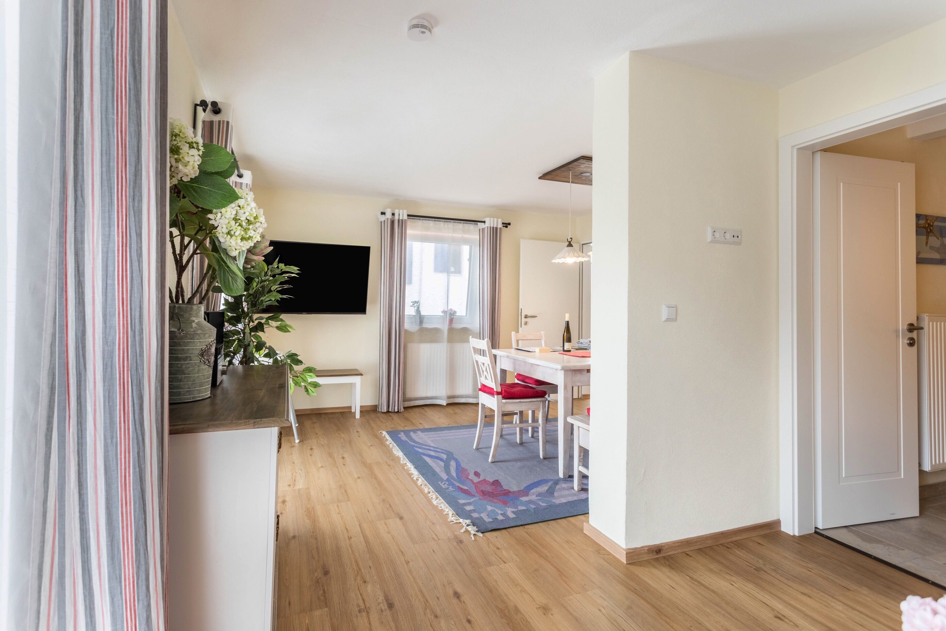 Ein Zimmer mit Holzboden, einem Esstisch mit Stühlen, einem an der Wand befestigten Fernseher und einer Tür zu einem anderen Raum.