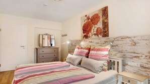 A bedroom with a bed, a nightstand, a dresser, a mirror, and a floral artwork on the wall.