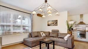 A living room with a sectional sofa, a small table, a chandelier, a kitchen area, and a large window with curtains.