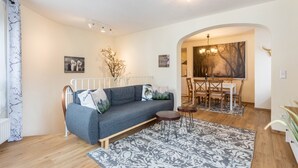 Uma sala de estar moderna com um sofá cinzento, uma mesa de jantar com cadeiras, um tapete estampado e um quadro na parede.