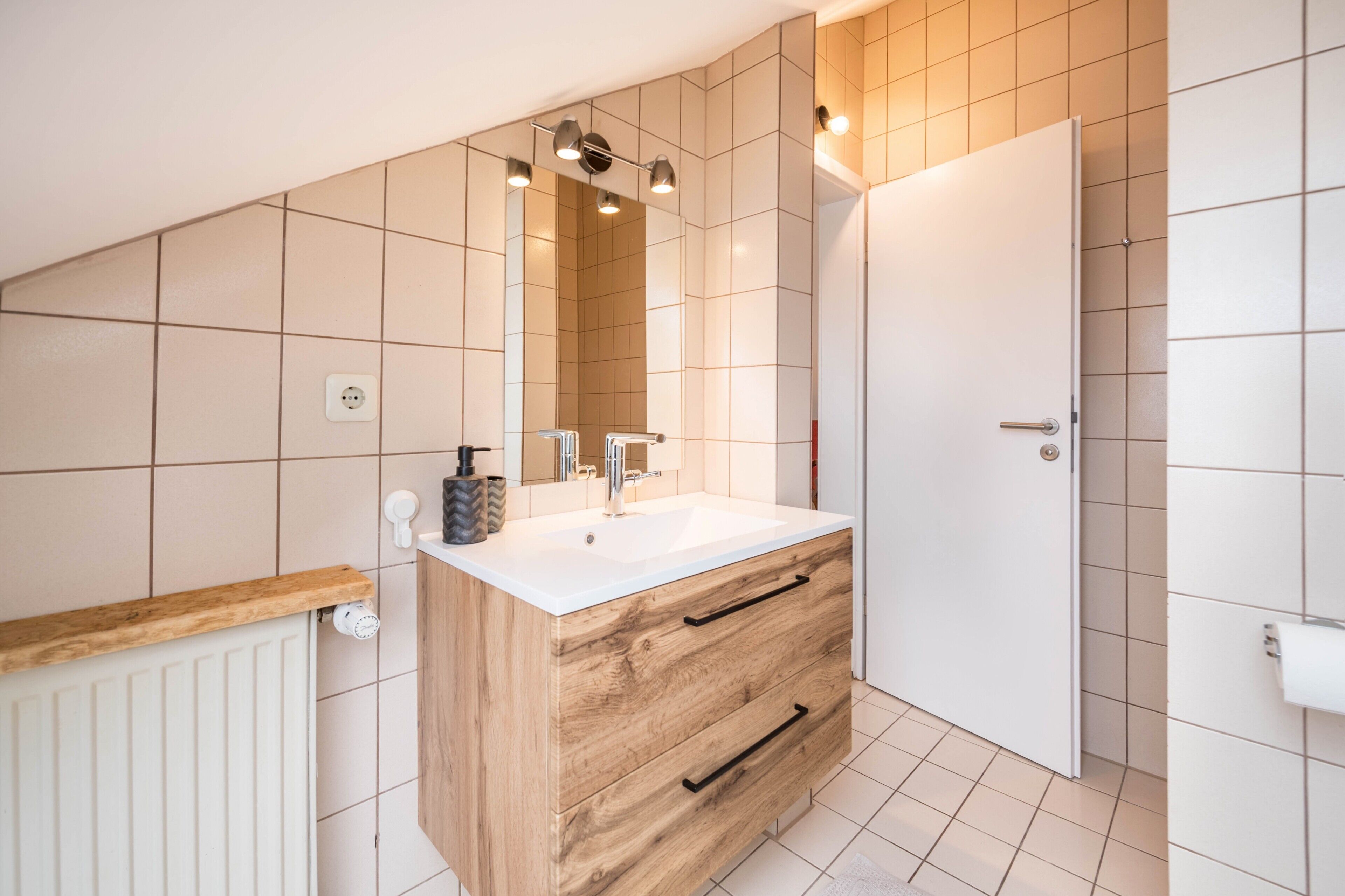 Un baño con un mueble de tocador de madera, un lavabo blanco y un espejo encima. Hay una puerta blanca y un radiador blanco en la pared.