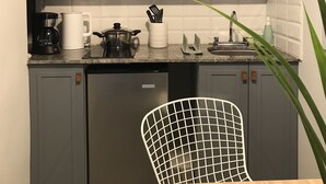 Eine kleine Küche mit grauen Schränken, einem integrierten Geschirrspüler, einem Holztisch und einem weißen Stuhl. Darüber befinden sich Regale mit Geschirr, Gläsern und Besteck.