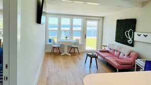 Un saló modern amb un sofà rosa, una taula rodona amb cadires i vista al mar a través de grans finestres.