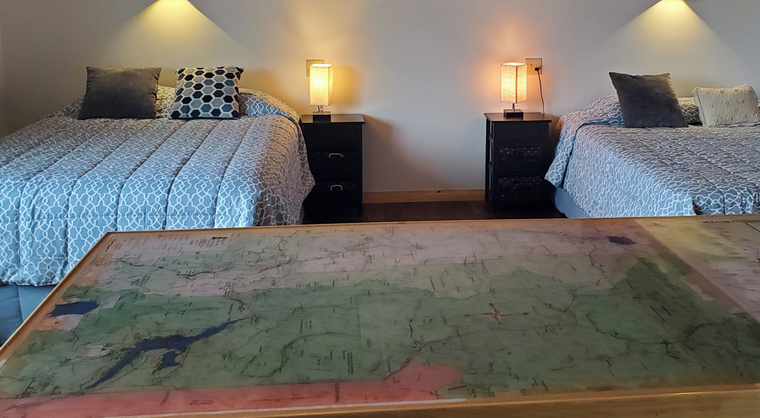 Quarto com duas camas, cada uma com roupa de cama estampada e uma mesa de cabeceira com abajur. Uma mesa grande e colorida está em primeiro plano.