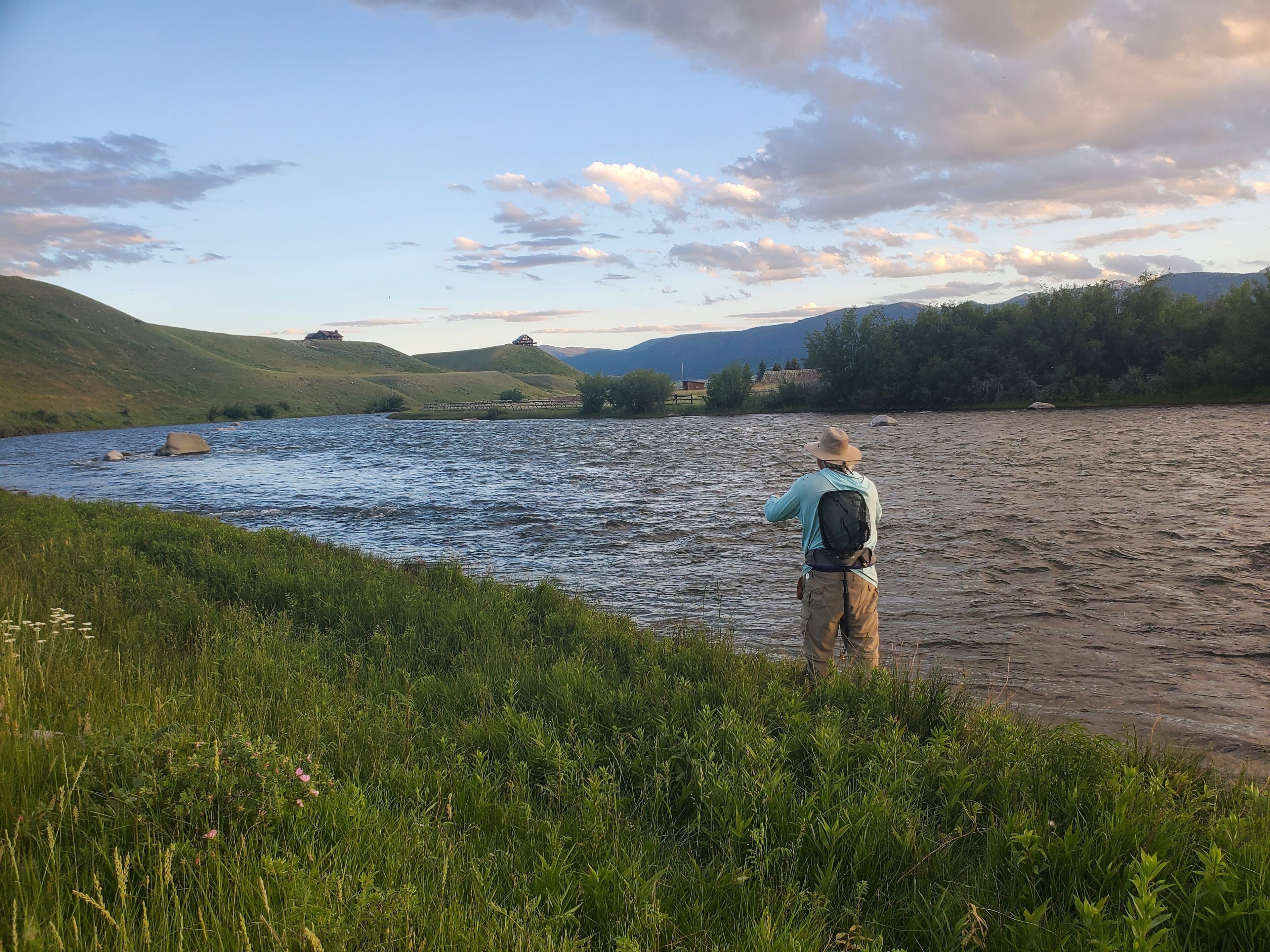 A person standing by a river with a backpack, surrounded by greenery and hills in the background.