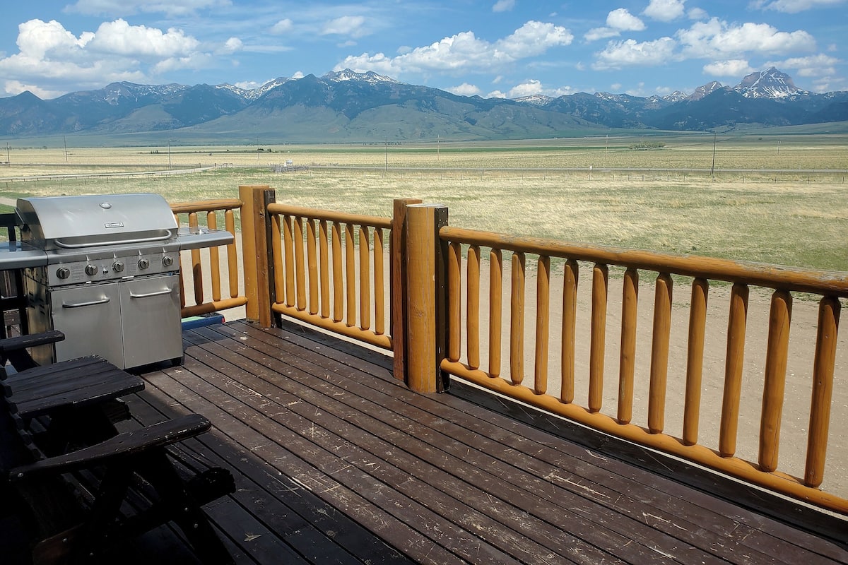 A deck with a grill and a table, overlooking a grassy field and mountains.
