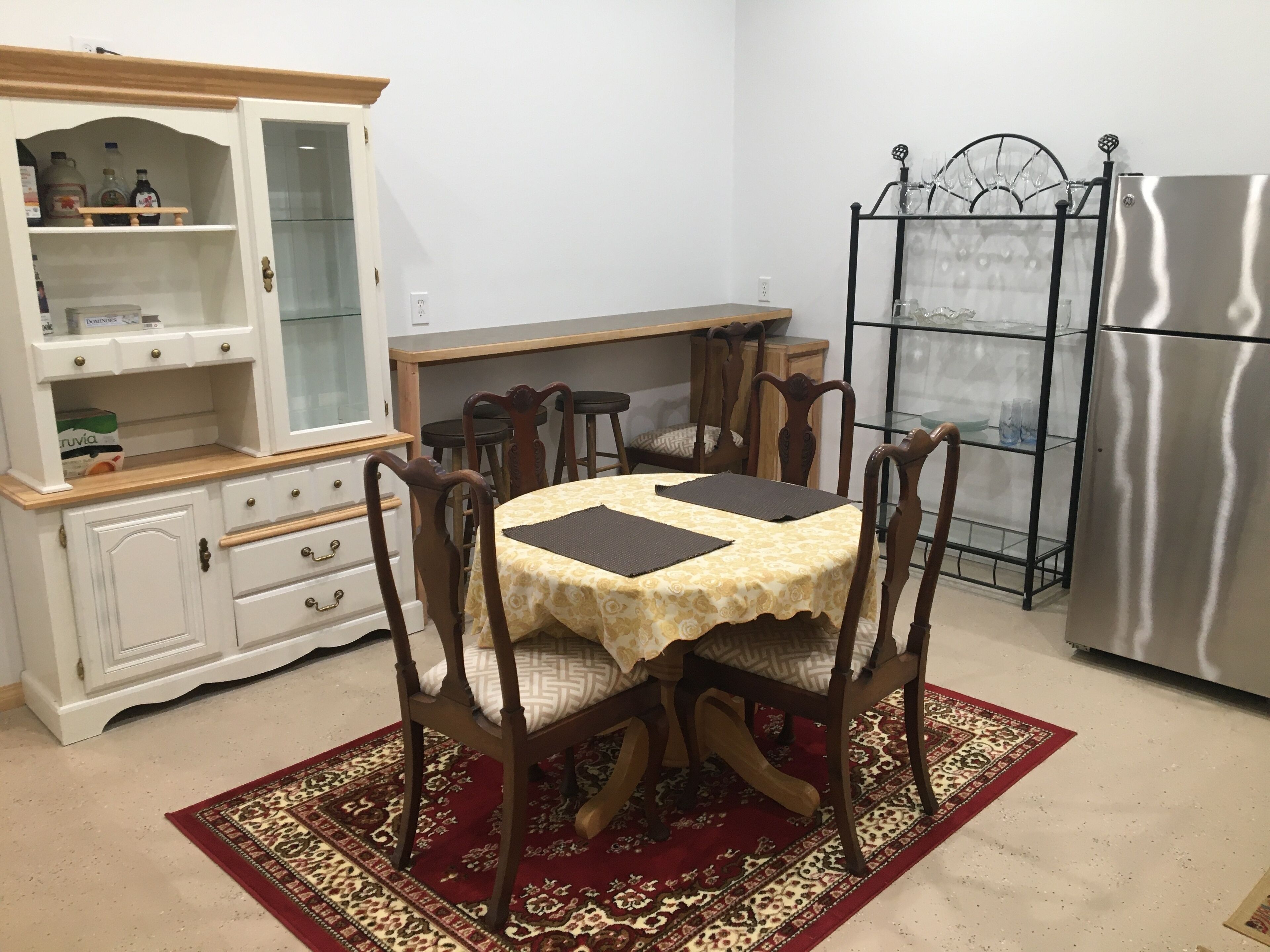 A dining area with a wooden table, chairs, a red patterned rug, a white cabinet with glass doors, and a stainless steel refrigerator.
