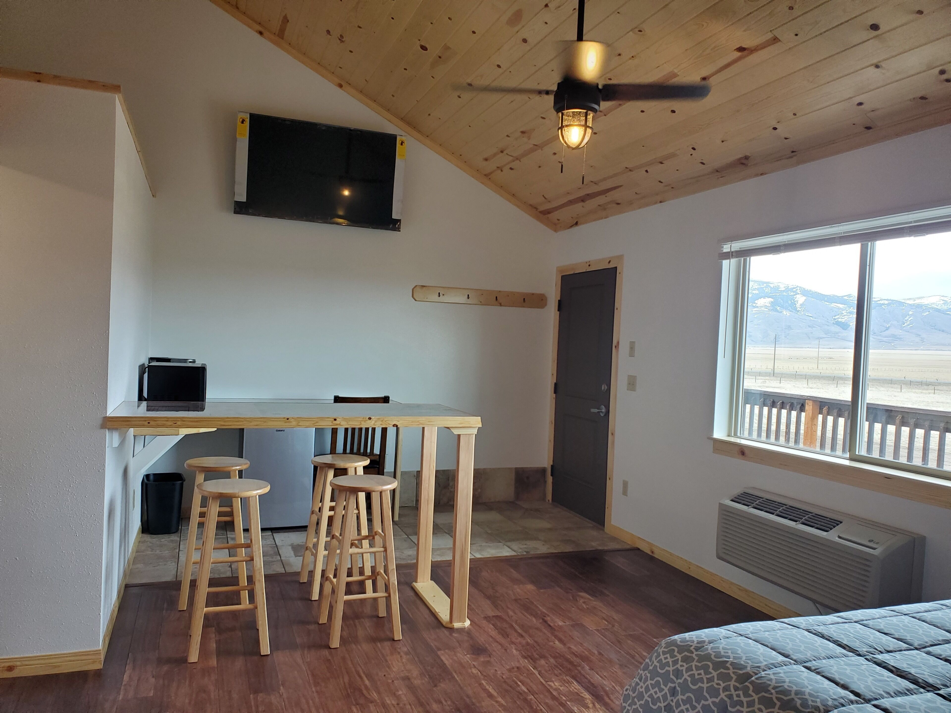 Une pièce avec un bureau en bois, deux tabourets, un téléviseur fixé au mur, une porte et une fenêtre donnant sur les montagnes.