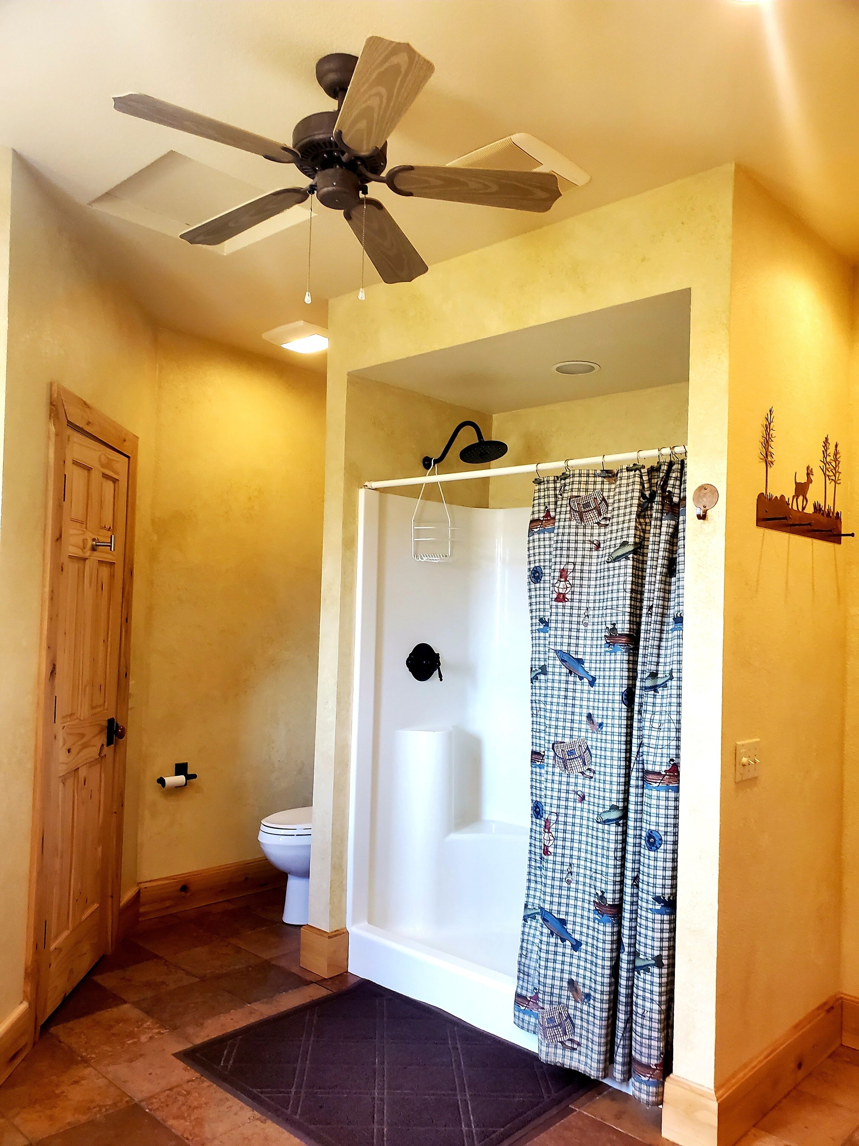 A bathroom with a ceiling fan, a shower, a toilet, and a patterned shower curtain.