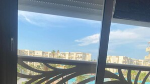 Vista desde una ventana con balcón, mostrando una piscina y edificios al fondo.