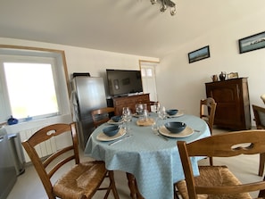 A dining area with a round table set for four, surrounded by wooden chairs. A refrigerator and a television are visible in the background.