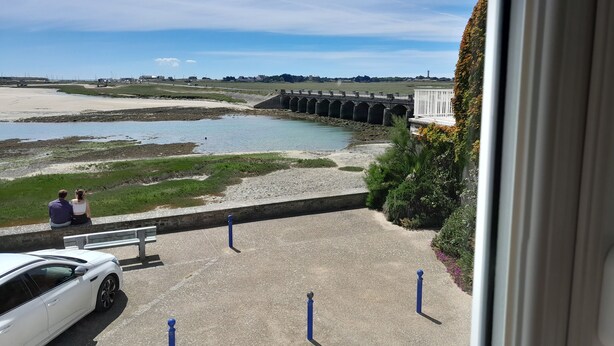 A view of a coastal area with a bridge, a white car parked in a driveway, and two people sitting on a bench.