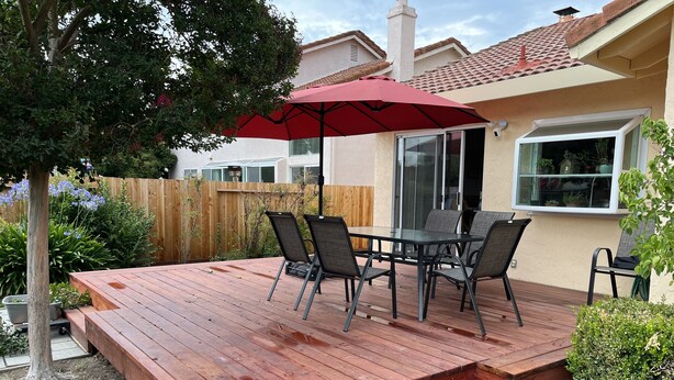 Una terraza de madera con una mesa, sillas, un paraguas rojo y una valla de madera al fondo.