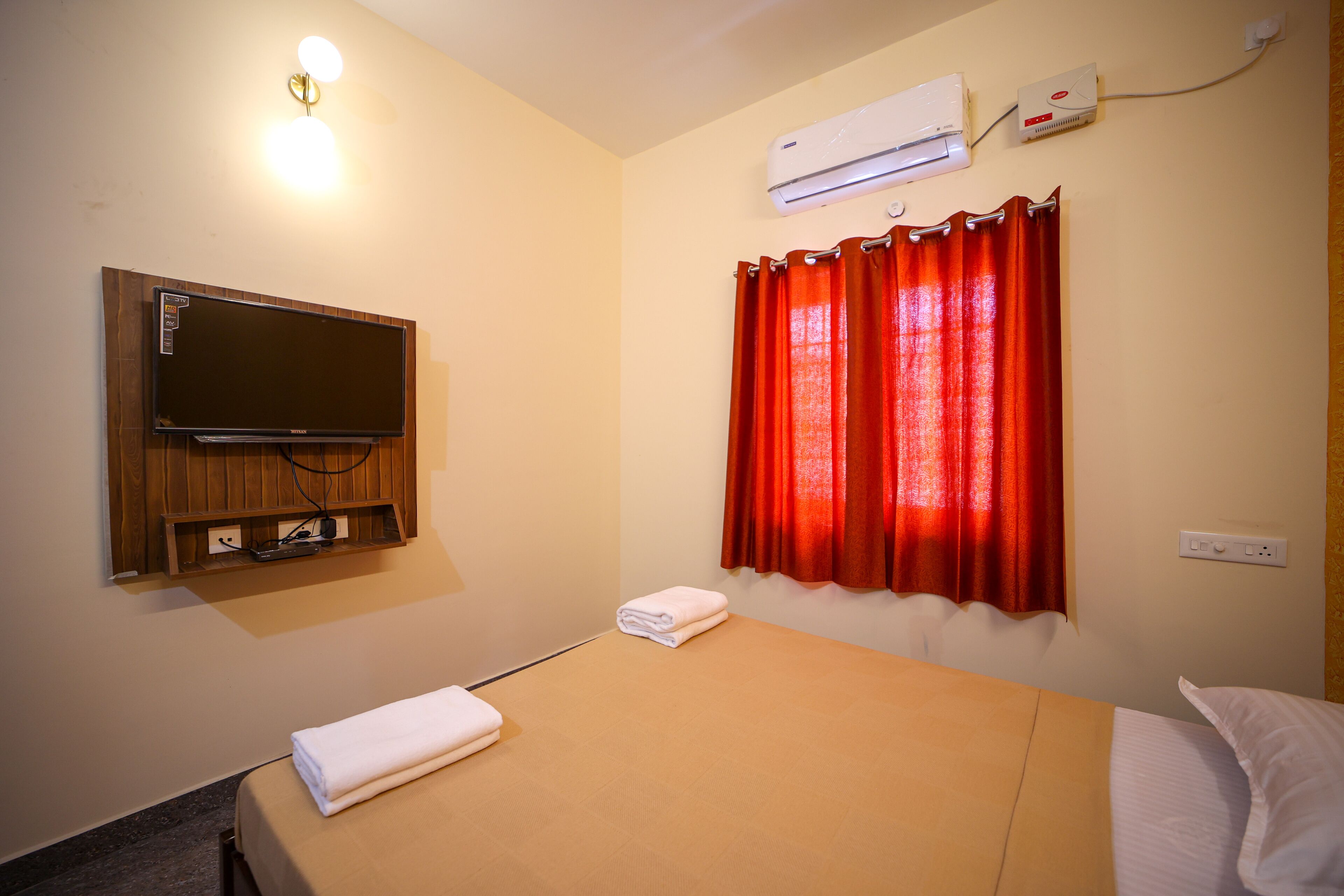 Une chambre avec un lit, un téléviseur fixé au mur, un appareil de climatisation et une fenêtre avec des rideaux rouges.