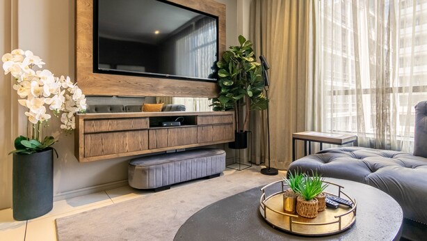 Een moderne woonkamer met een grote flatscreen televisie aan een houten wand, een grijze bank, een ronde salontafel met decoratieve objecten en een potplant.