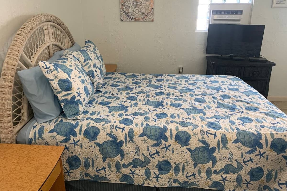 A bedroom with a bed, a wicker chair, a television, and a wall-mounted air conditioner.