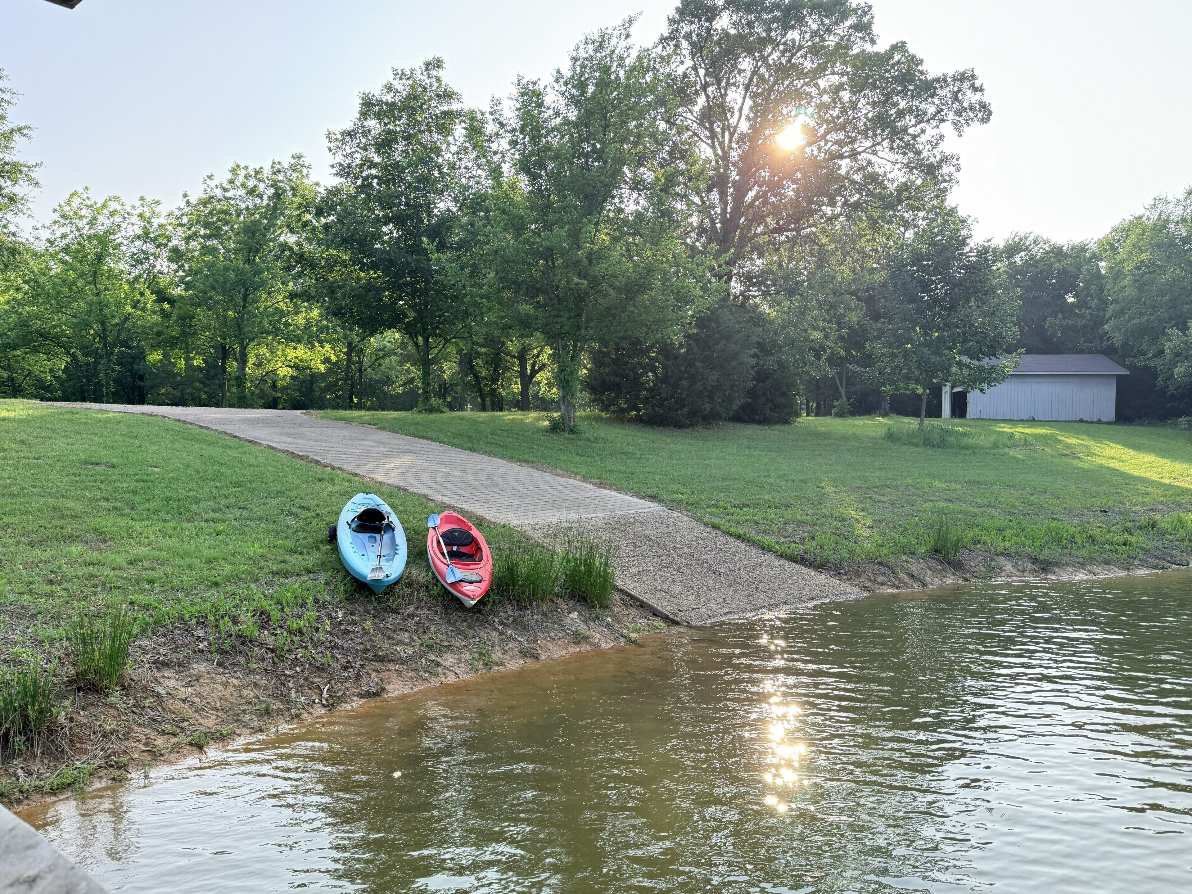 A grassy area with a small bridge leading to a body of water, kayaks, and a shed in the background.