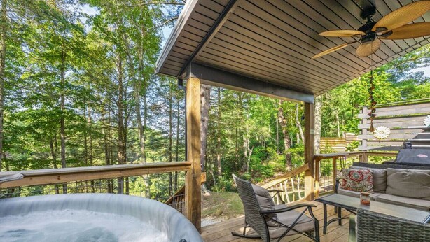 A hot tub on a wooden deck with a view of a forest. There is a rocking chair and a table with cushions. - Hemlocks Over Muir-WiFi, Firepit, Hot Tub, located in Muir Valley (Rogers)