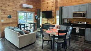 A cozy cabin interior with a sofa, dining table, chairs, a kitchen area with appliances, and a large window. - Hemlocks Over Muir-WiFi, Firepit, Hot Tub, located in Muir Valley (Rogers)
