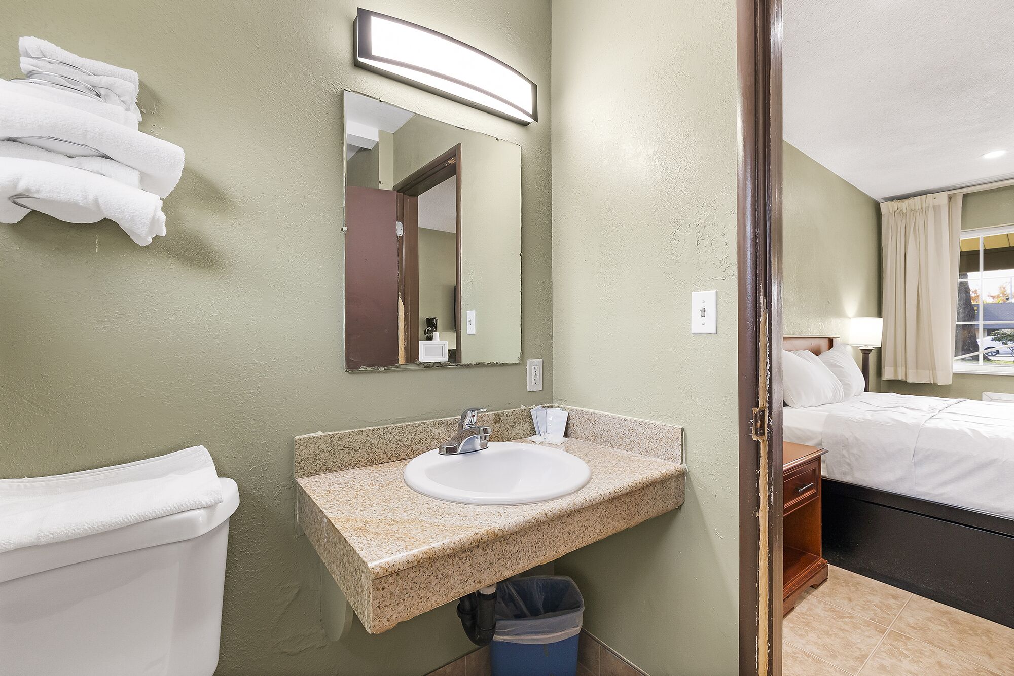 Bagno d'hotel con lavabo su piano in granito, specchio, servizi igienici e letto visibile attraverso una porta aperta.
