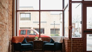 Ein Zimmer mit zwei Stühlen, einem kleinen Tisch und einem Fenster mit Blick auf ein Backsteingebäude und parkende Autos.